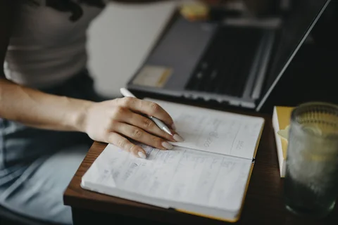 Hands writing in a notebook next to a laptop